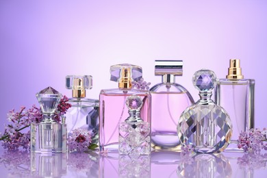 Photo of Bottles of perfume and lilac flowers on mirror surface against violet gradient background