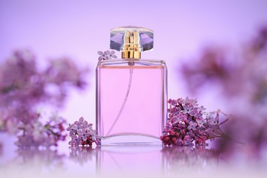 Photo of Bottle of perfume and lilac flowers on mirror surface against violet gradient background, selective focus