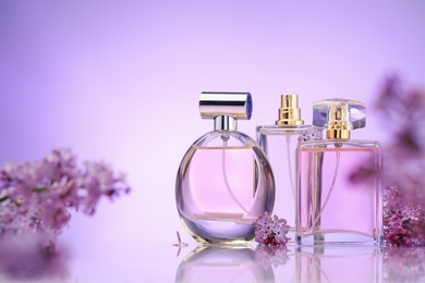 Photo of Bottles of perfume and lilac flowers on mirror surface against violet gradient background, selective focus