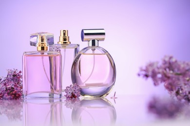 Photo of Bottles of perfume and lilac flowers on mirror surface against violet gradient background, selective focus