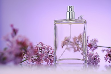 Photo of Bottle of perfume and lilac flowers on mirror surface against violet gradient background, selective focus