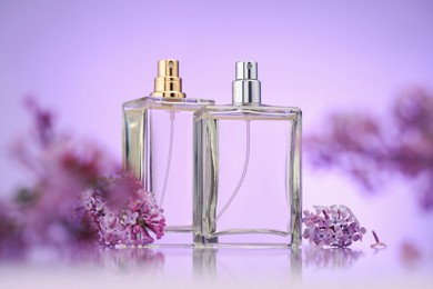 Photo of Bottles of perfume and lilac flowers on mirror surface against violet gradient background, selective focus