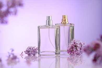Photo of Bottles of perfume and lilac flowers on mirror surface against violet gradient background, selective focus