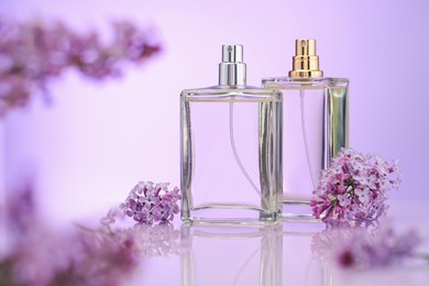 Photo of Bottles of perfume and lilac flowers on mirror surface against violet gradient background, selective focus
