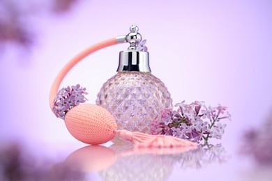 Photo of Bottle of perfume and lilac flowers on mirror surface against violet gradient background, selective focus