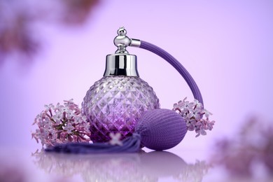 Photo of Bottle of perfume and lilac flowers on mirror surface against violet gradient background, selective focus