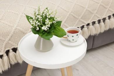 Photo of Bouquet of beautiful lily-of-the-valley in vase and cup of tea on coffee table indoors