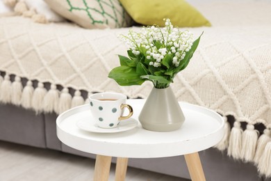 Photo of Bouquet of beautiful lily-of-the-valley in vase and cup of tea on coffee table indoors