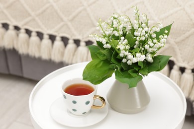 Photo of Bouquet of beautiful lily-of-the-valley in vase and cup of tea on coffee table indoors
