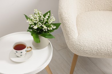 Photo of Bouquet of beautiful lily-of-the-valley in vase and cup of tea on coffee table near armchair indoors