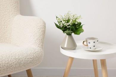 Photo of Bouquet of beautiful lily-of-the-valley in vase and cup of tea on coffee table near armchair indoors