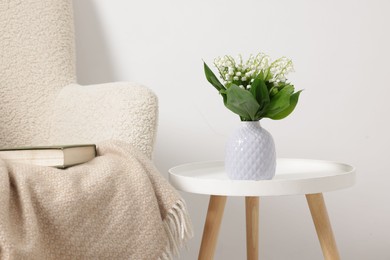 Photo of Bouquet of beautiful lily-of-the-valley in vase on coffee table near armchair indoors