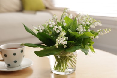 Photo of Bouquet of beautiful lily-of-the-valley and cup on table indoors, closeup