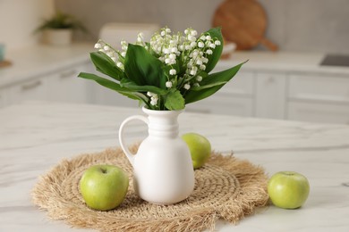 Photo of Bouquet of beautiful lily-of-the-valley in vase and apples on white marble table in kitchen, closeup
