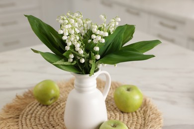 Photo of Bouquet of beautiful lily-of-the-valley in vase and apples on white marble table in kitchen, closeup