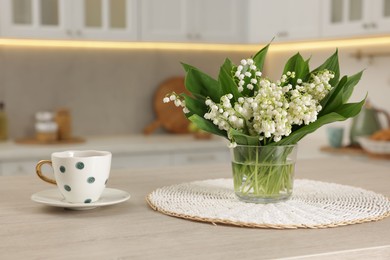 Photo of Bouquet of beautiful lily-of-the-valley and cup on wooden table in kitchen