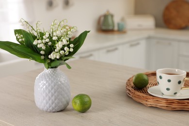 Photo of Beautiful lily-of-the-valley in vase, limes and cup of tea on wooden table in kitchen, closeup