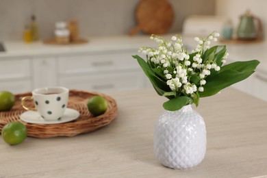 Photo of Beautiful lily-of-the-valley in vase, limes and cup of tea on wooden table in kitchen, closeup