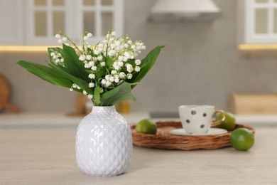 Photo of Beautiful lily-of-the-valley in vase, limes and cup of tea on wooden table in kitchen, closeup