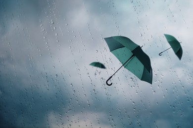 Image of Dark green umbrellas flying in cloudy sky, view through wet window. Stormy weather