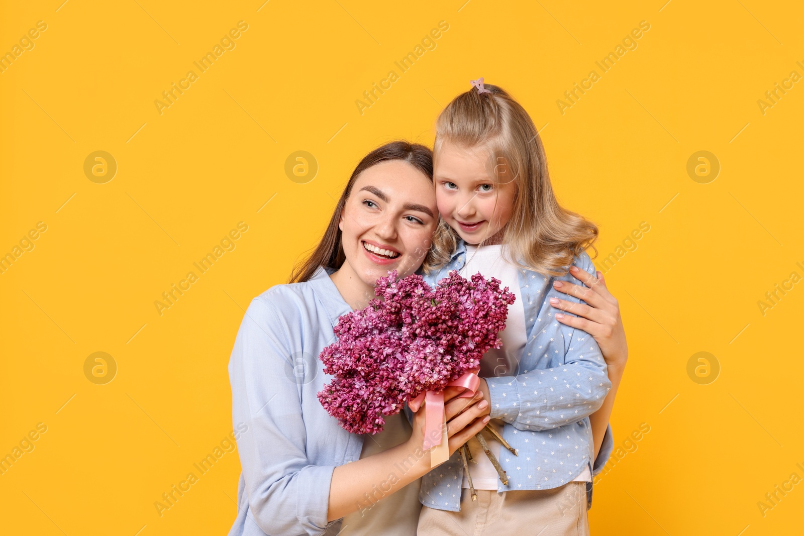 Mother and her daughter with bouquet of lilac flowers on yellow background Photo of Mother and her daughter with bouquet of lilac flowers on yellow background