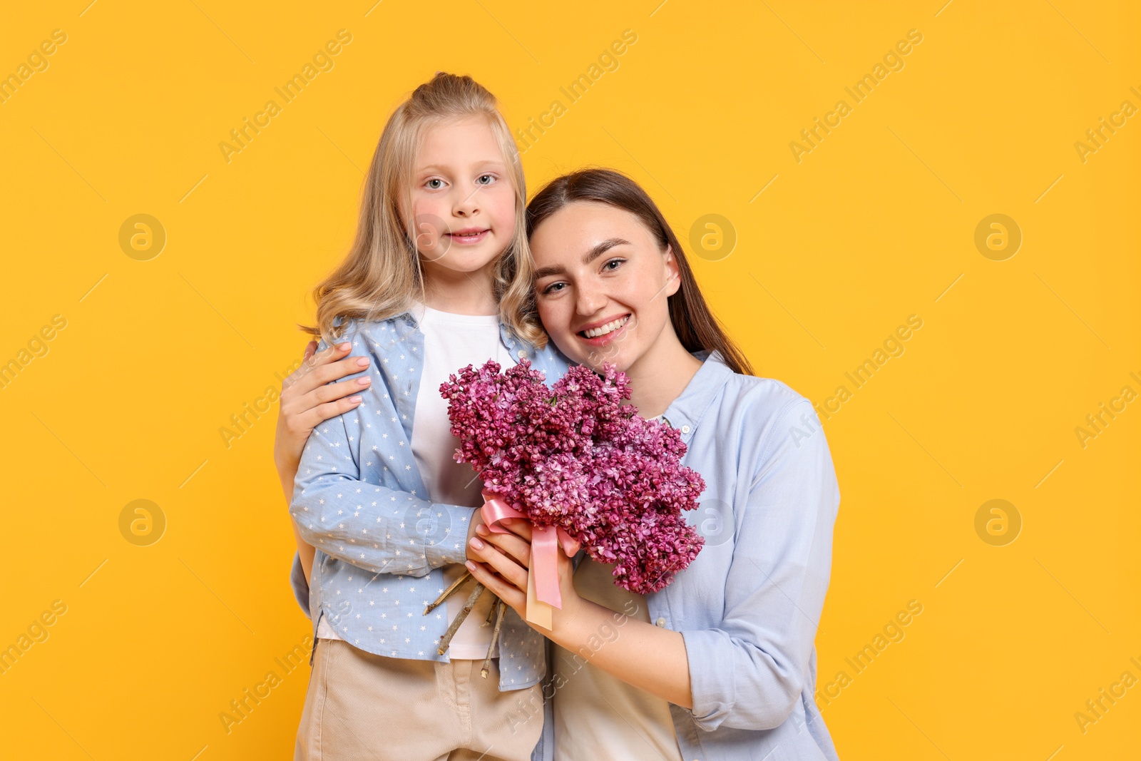 Mother and her daughter with bouquet of lilac flowers on yellow background Photo of Mother and her daughter with bouquet of lilac flowers on yellow background