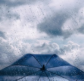 Image of Blue umbrella under cloudy sky, view through wet window. Stormy weather