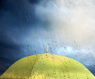 Image of Yellow umbrella under cloudy sky, view through wet window. Stormy weather