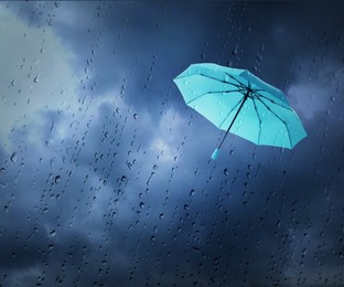 Image of Light blue umbrella flying in dark cloudy sky, view through wet window. Stormy weather