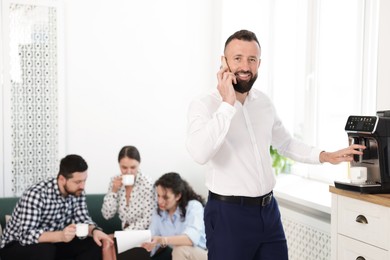Photo of Man talking on phone while making coffee. Colleagues having break in office