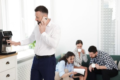 Photo of Man talking on phone while making coffee. Colleagues having break in office