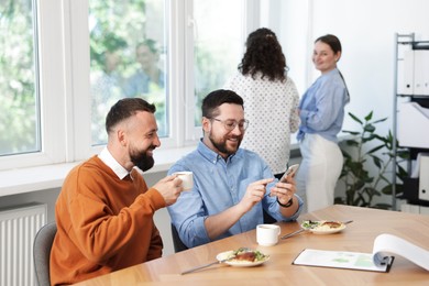 Photo of Man showing his colleague something on smartphone during lunch break in office