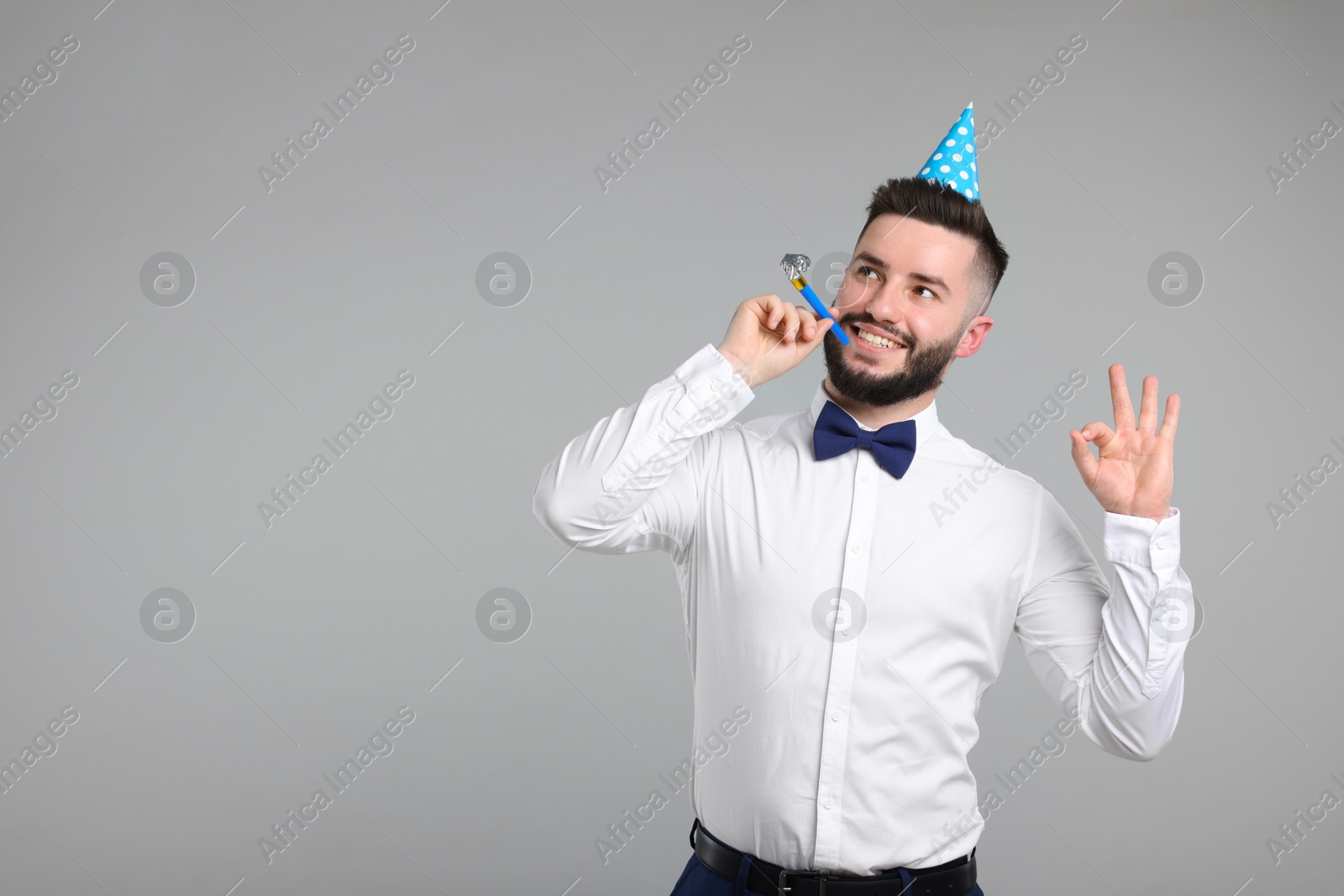 Photo of Happy Birthday. Handsome man with party hat and blower showing ok gesture on grey background, space for text
