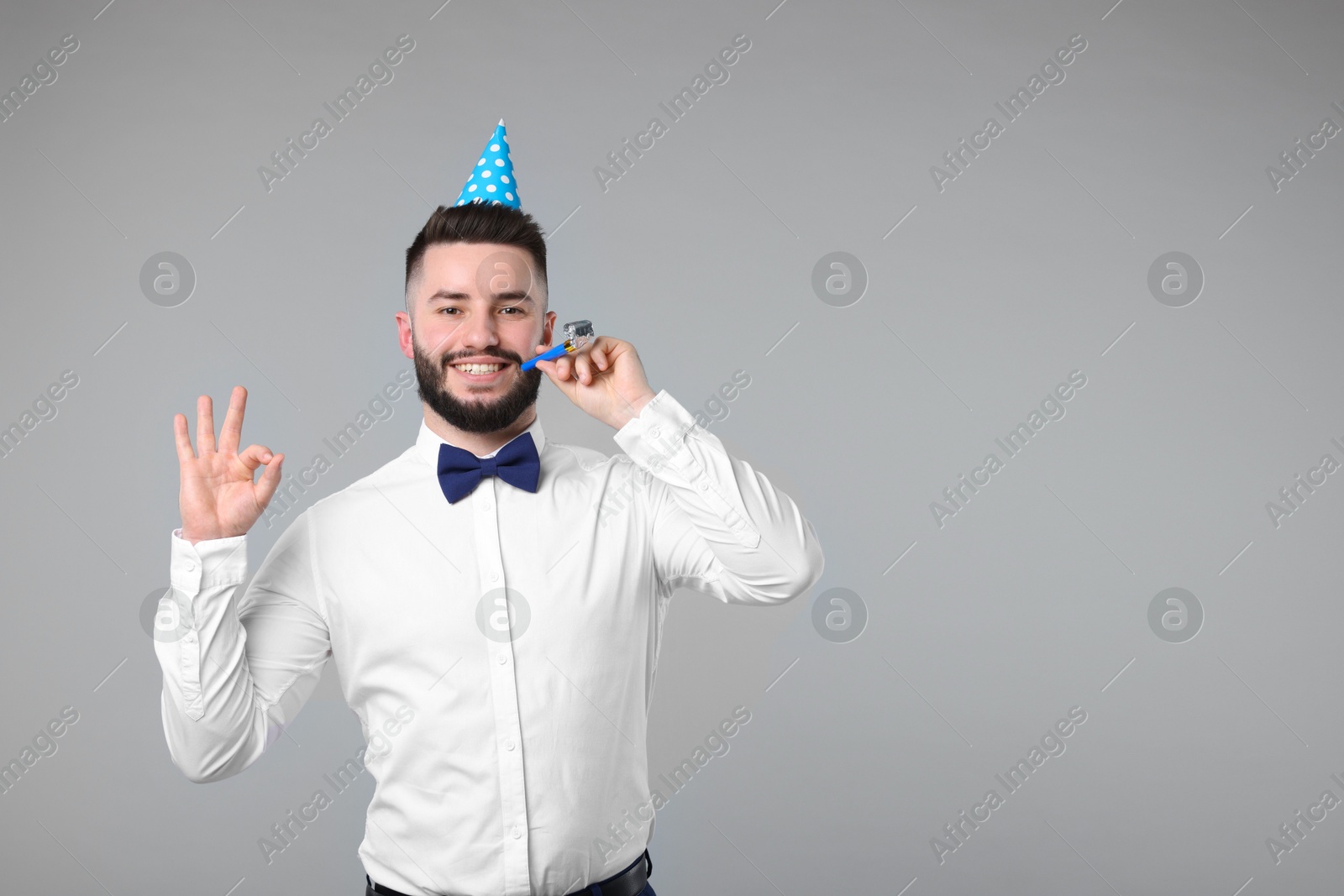 Happy Birthday. Handsome man with party hat and blower showing ok gesture on grey background, space for text Photo of Happy Birthday. Handsome man with party hat and blower showing ok gesture on grey background, space for text