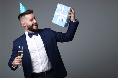 Photo of Happy Birthday. Handsome man with party hat, gift box and glass of champagne on grey background, space for text