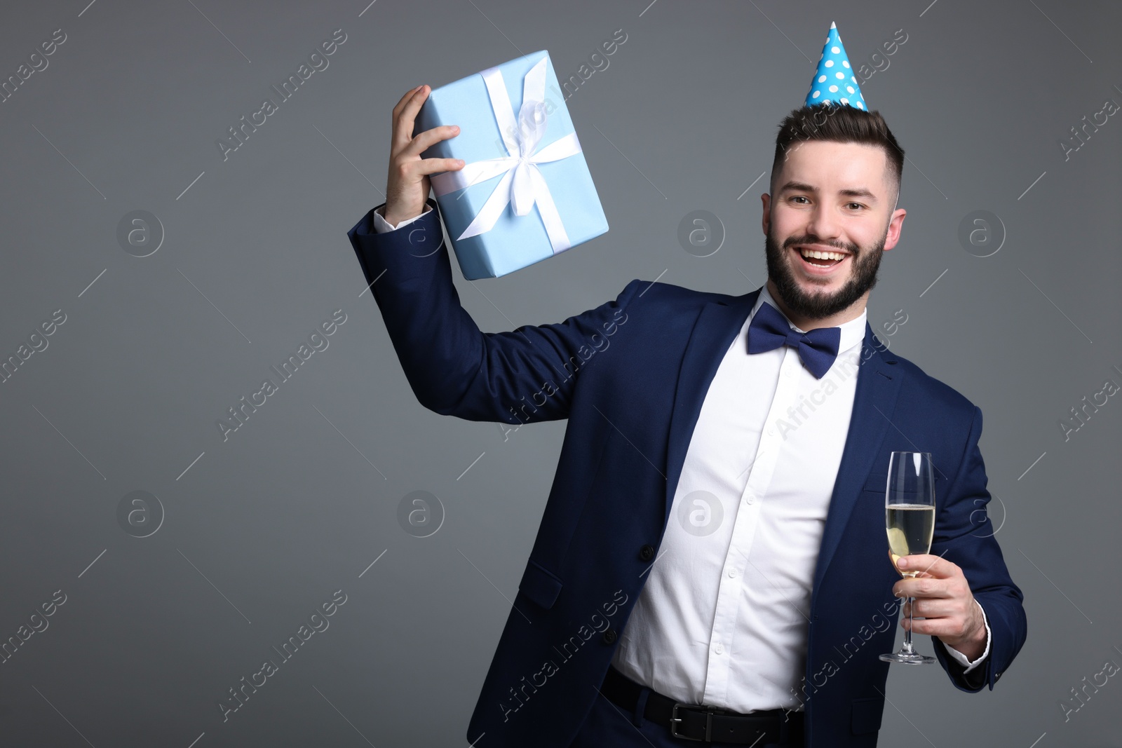 Happy Birthday. Handsome man with party hat, gift box and glass of champagne on grey background, space for text Photo of Happy Birthday. Handsome man with party hat, gift box and glass of champagne on grey background, space for text