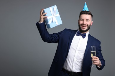 Photo of Happy Birthday. Handsome man with party hat, gift box and glass of champagne on grey background, space for text