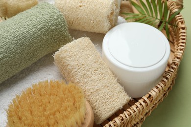 Photo of Natural loofah sponges, cosmetic product, towels, brush and twig in basket on green background, closeup