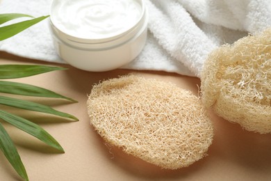 Photo of Natural loofah sponges, cosmetic product, towel and green leaves on beige background, closeup