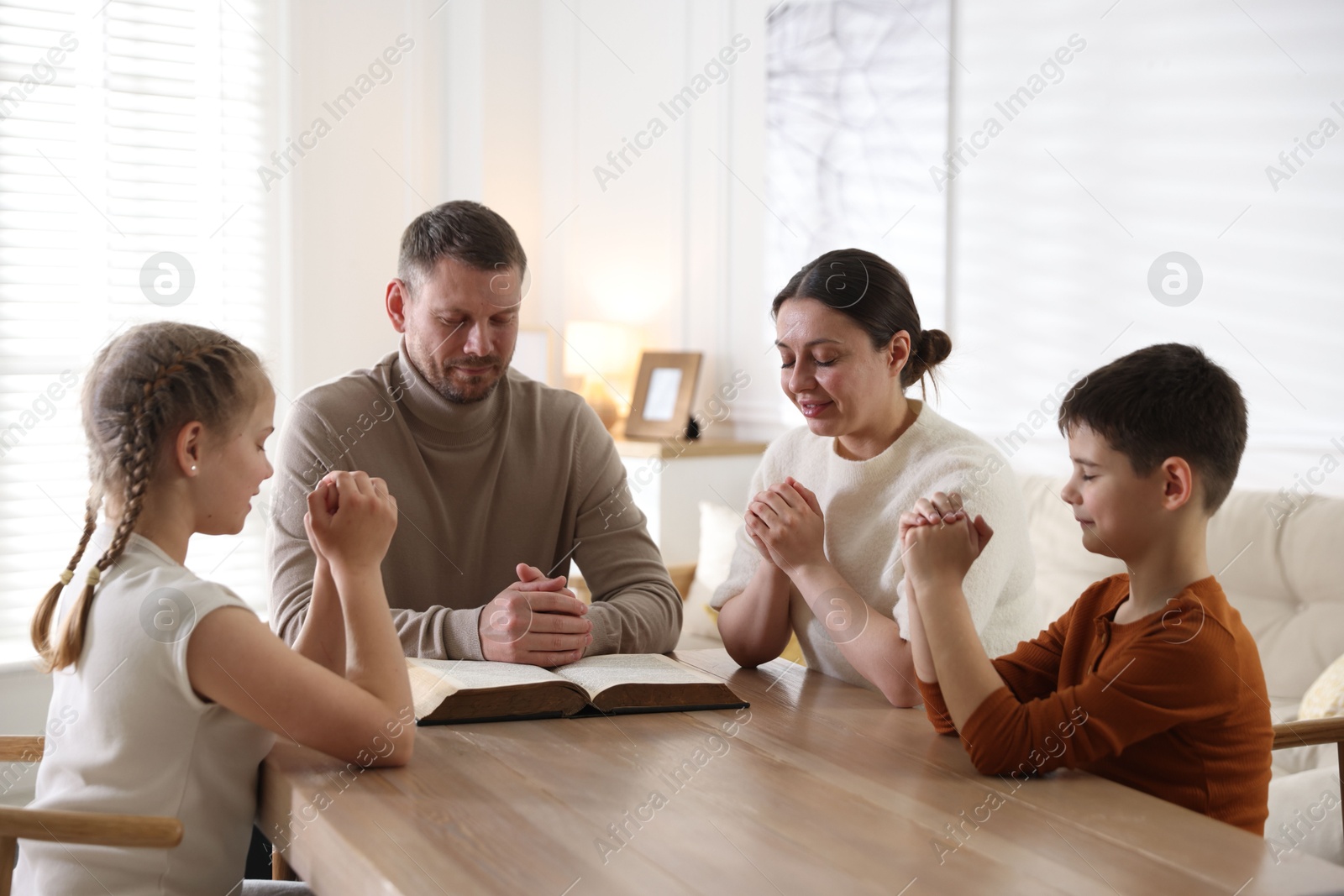 Family with Bible praying at table indoors Photo of Family with Bible praying at table indoors