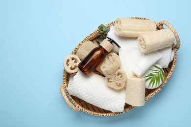 Photo of Natural loofah sponges, towels, cosmetic products and floral decor in basket on light blue background, top view. Space for text