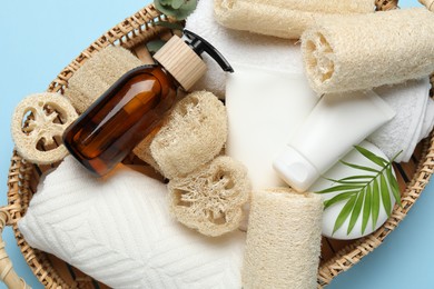 Photo of Natural loofah sponges, towels, cosmetic products and floral decor in basket on light blue background, top view
