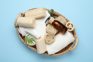 Photo of Natural loofah sponges, towels, cosmetic products and floral decor in basket on light blue background, top view