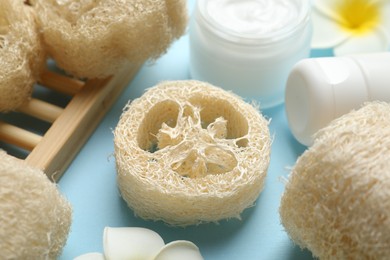 Photo of Natural loofah sponges, cosmetic products and plumeria flowers on light blue background, closeup