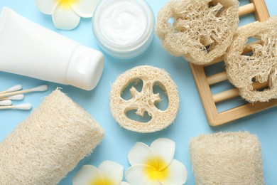 Photo of Natural loofah sponges, cosmetic products, swabs and plumeria flowers on light blue background, flat lay