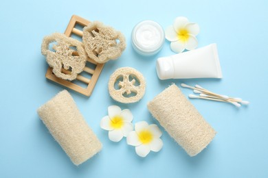Photo of Natural loofah sponges, cosmetic products, swabs and plumeria flowers on light blue background, flat lay