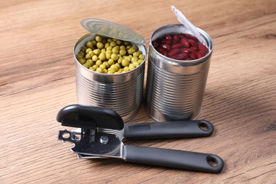 Photo of Can opener and tins with products on wooden table, closeup