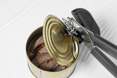 Photo of Can opener and tin with fish on white wooden table, above view