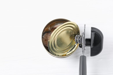 Photo of Can opener and tin with fish on white wooden table, top view. Space for text