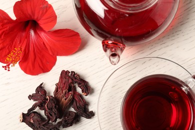 Photo of Delicious hibiscus tea in glass cup, teapot, dry roselle sepals and flower on light wooden table, flat lay